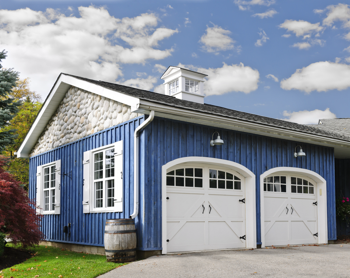 A detached 2-car garage with blue siding, white doors and window shutters. The roof is topped with a white cupola.