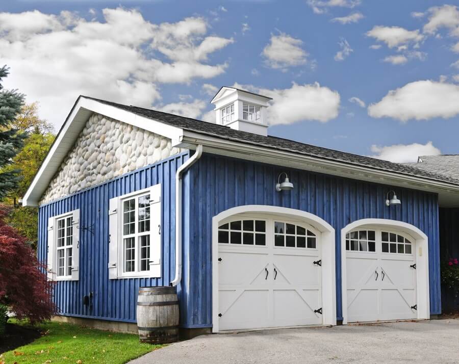 A detached 2-car garage with blue siding, white doors and window shutters. The roof is topped with a white cupola.